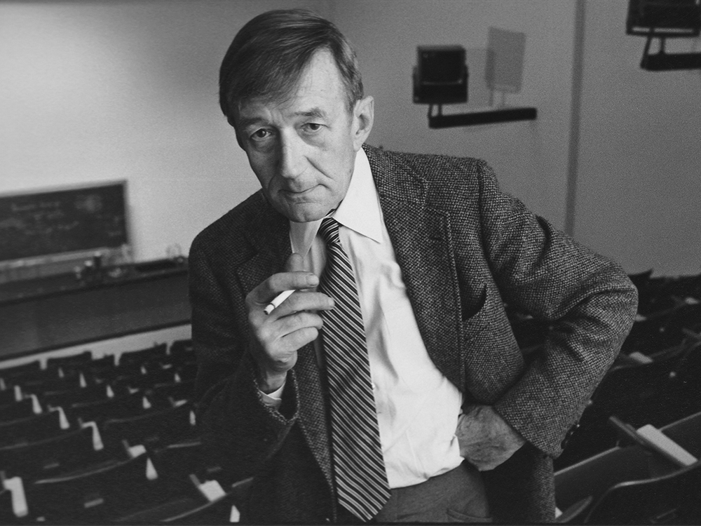 Black and white photo of a man posing for the camera with one hand on his hip and the other hand holding a cigarette